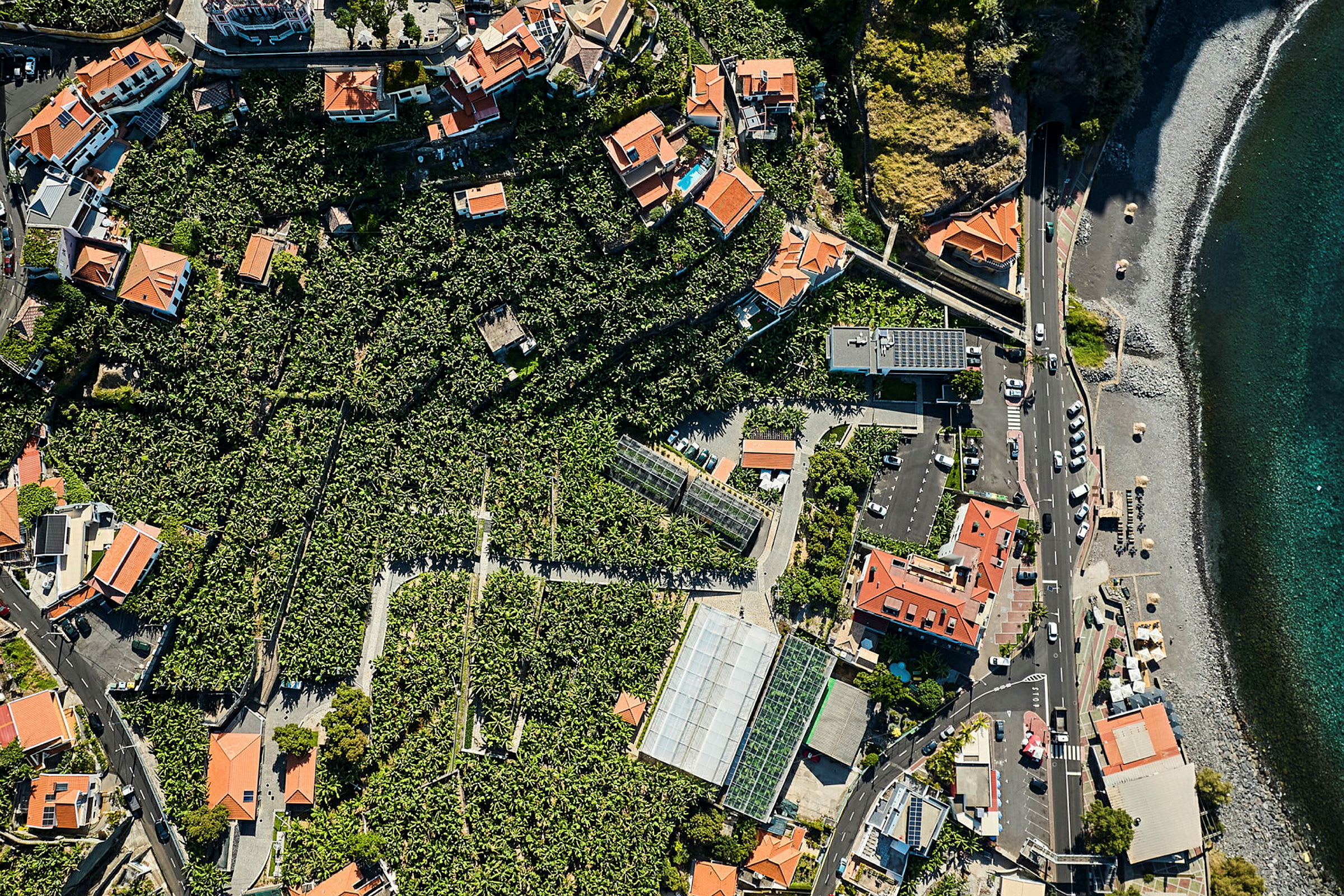 Luftfoto af et område med huse med røde tage, omgivet af grøn vegetation og tæt på en strand med flere biler parkeret langs en vej. (AI-genereret tekst)