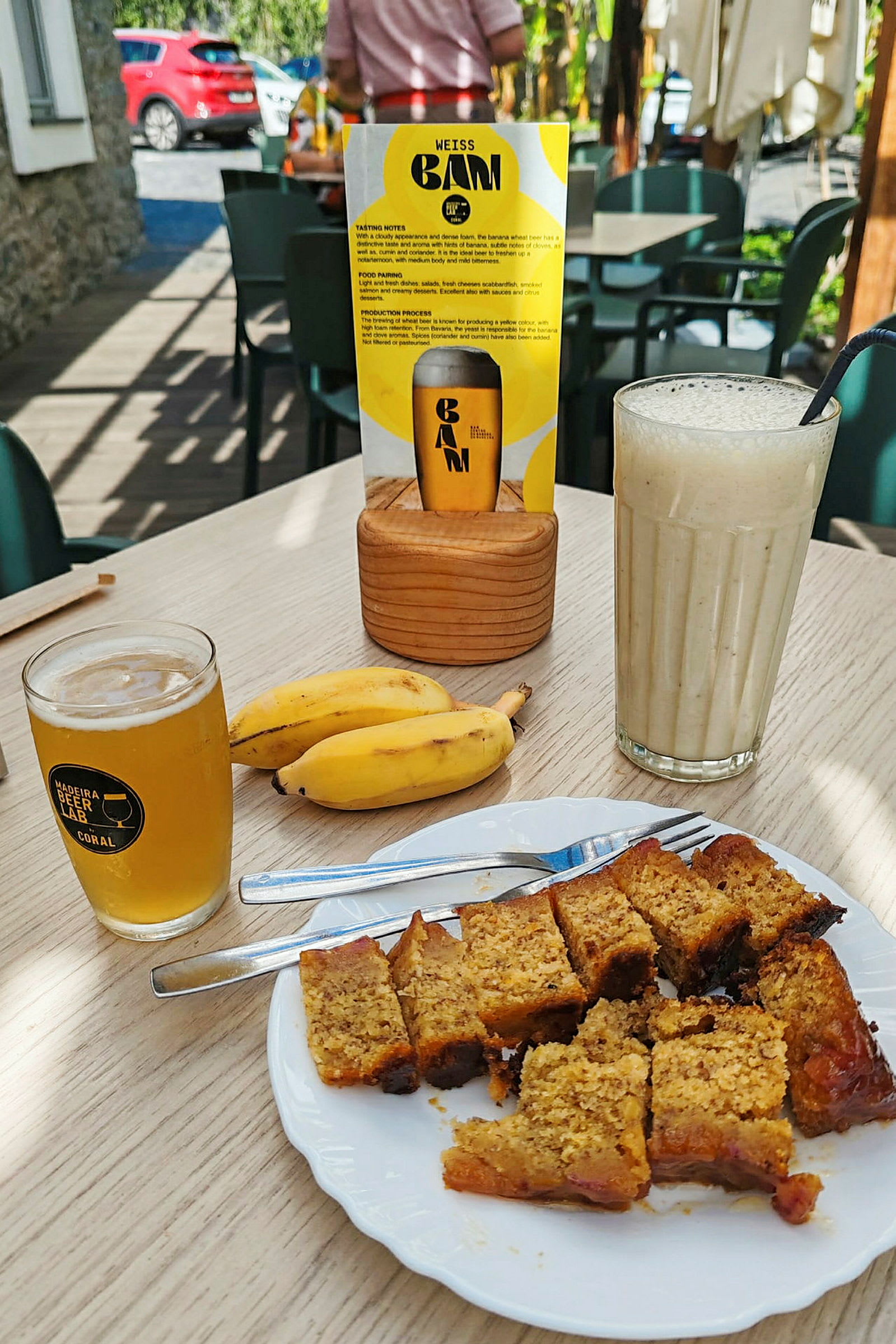 En tallerken med kagestykker, to bananer, et glas øl og en milkshake på et bord. Bagved ses en menu og udendørs siddepladser. (AI-genereret tekst)