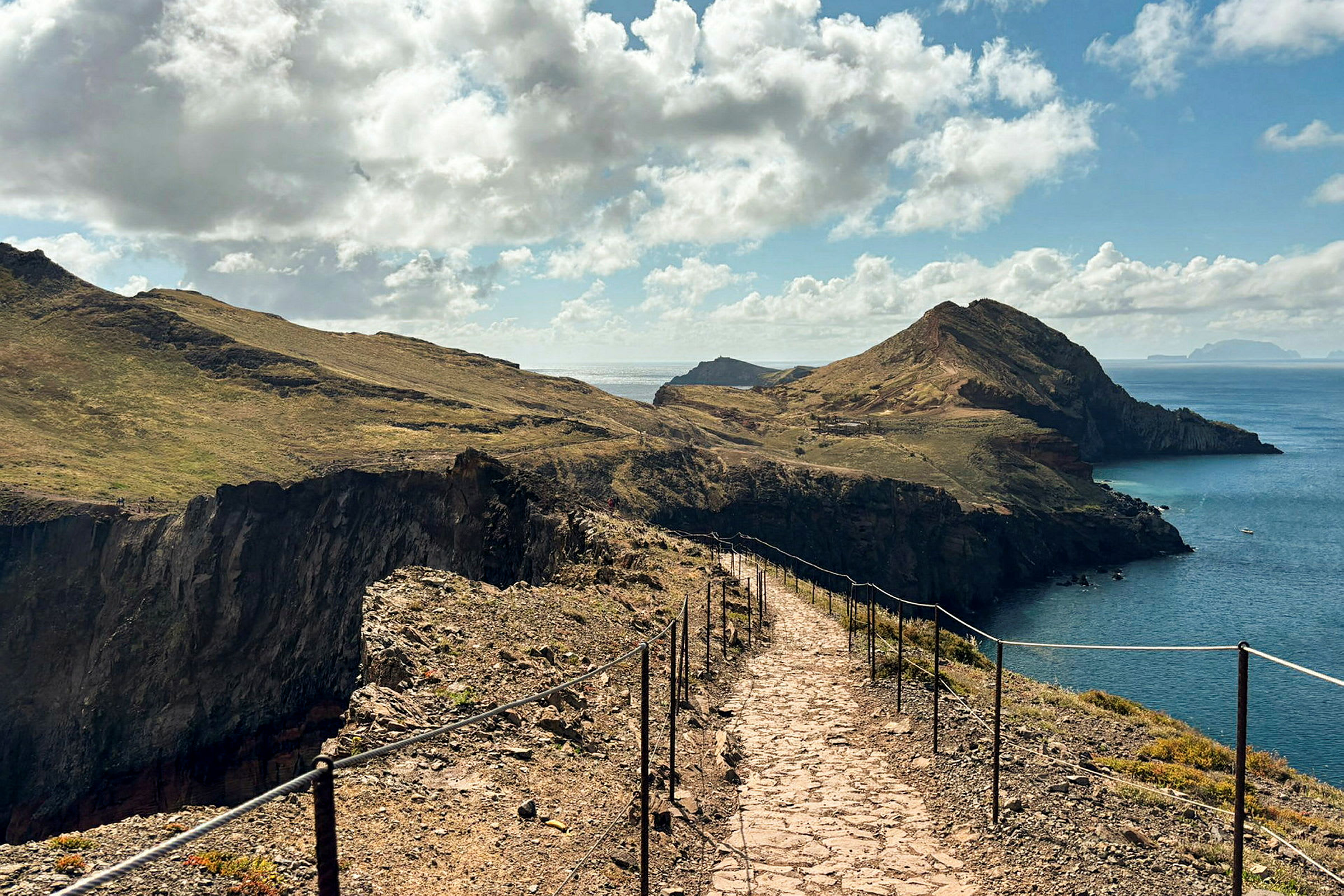 Et landskabsmotiv med en stenbelagt sti med rækværk på begge sider. Stien går langs en klippeskrænt omgivet af vegetation og leder mod bjerge nær kysten med havet i baggrunden. (AI-genereret tekst)
