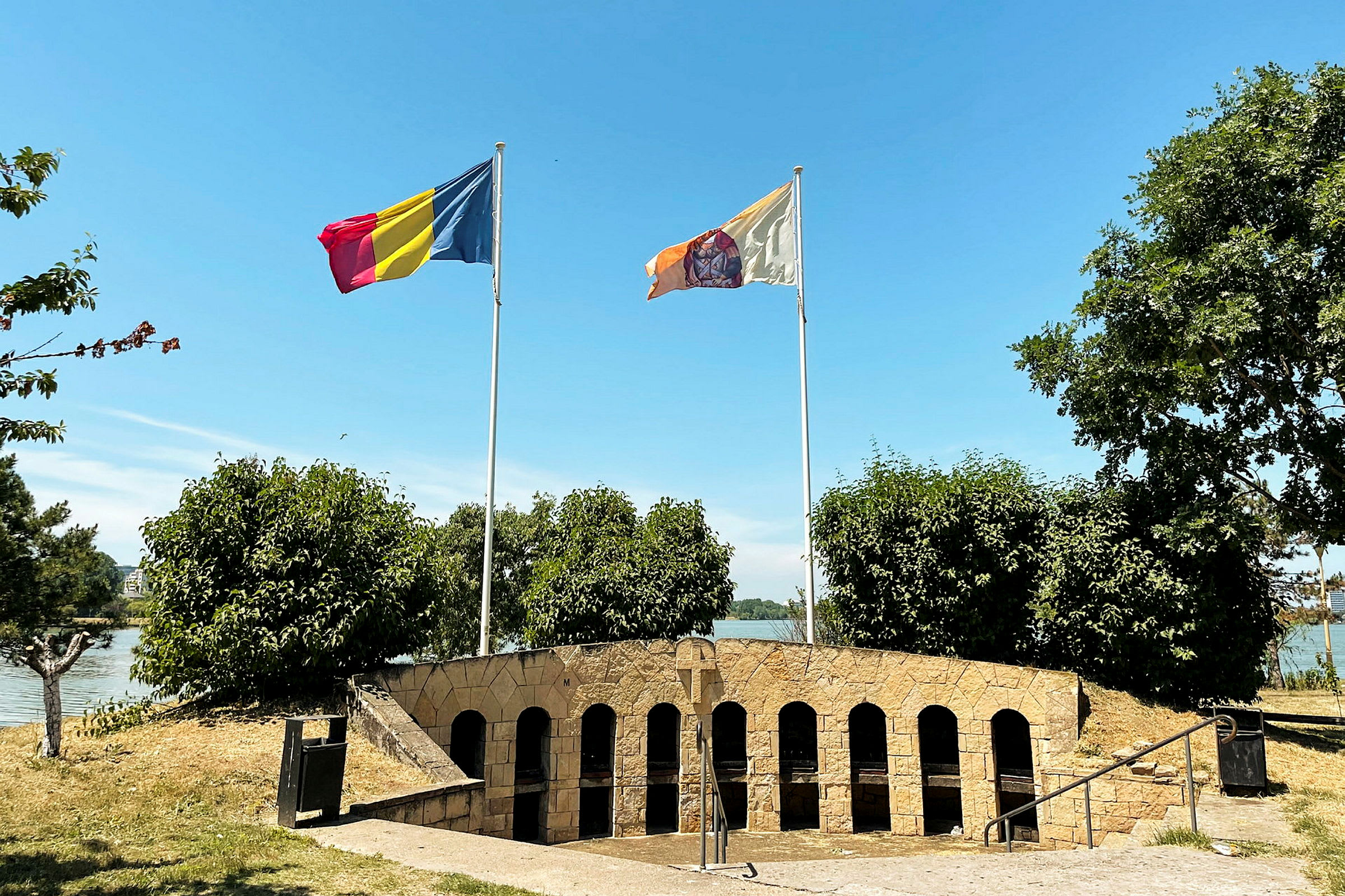 To flag vajende på flagstænger foran en stenstruktur med buede åbninger. Grønne buske og træer omgiver området, og en vandoverflade ses i baggrunden. (AI-genereret tekst)