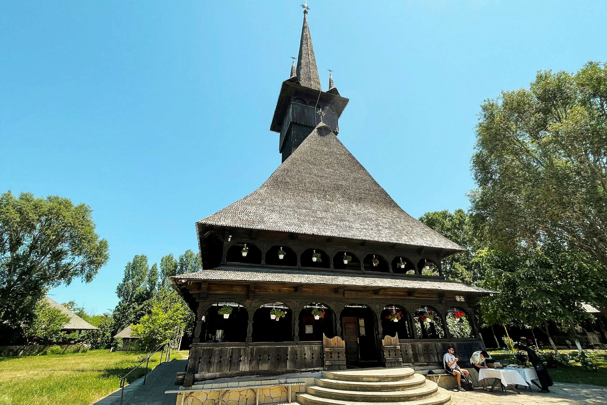 Nærbillede af en trækirke med spidst tag og dekorative elementer, omgivet af grønne områder og mennesker. (AI-genereret tekst)