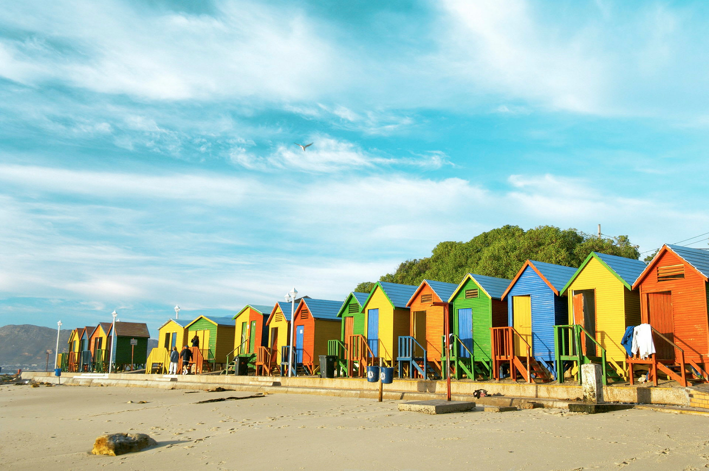 Flere farverige strandhuse står på række langs en sandstrand under en blå himmel. (AI-genereret tekst)