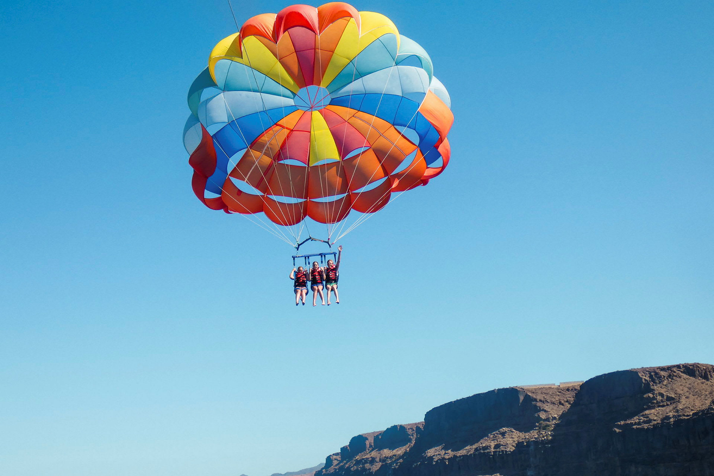 Tre personer som parasailer med en stor, farverig faldskærm mod en blå himmel. (AI-genereret tekst)