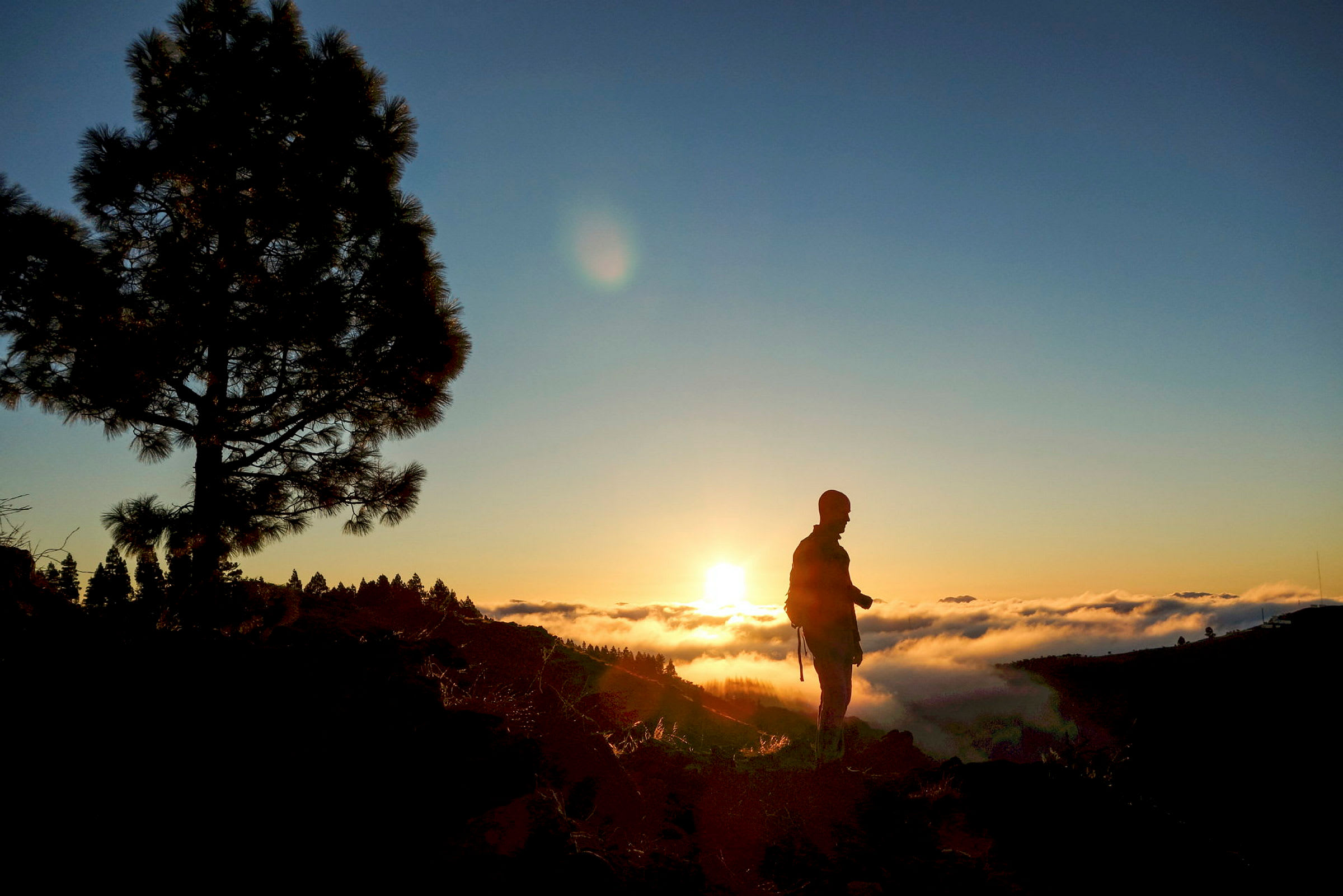 En person står på en høj ved solnedgang med skyer i dalen nedenfor. (AI-genereret tekst)
