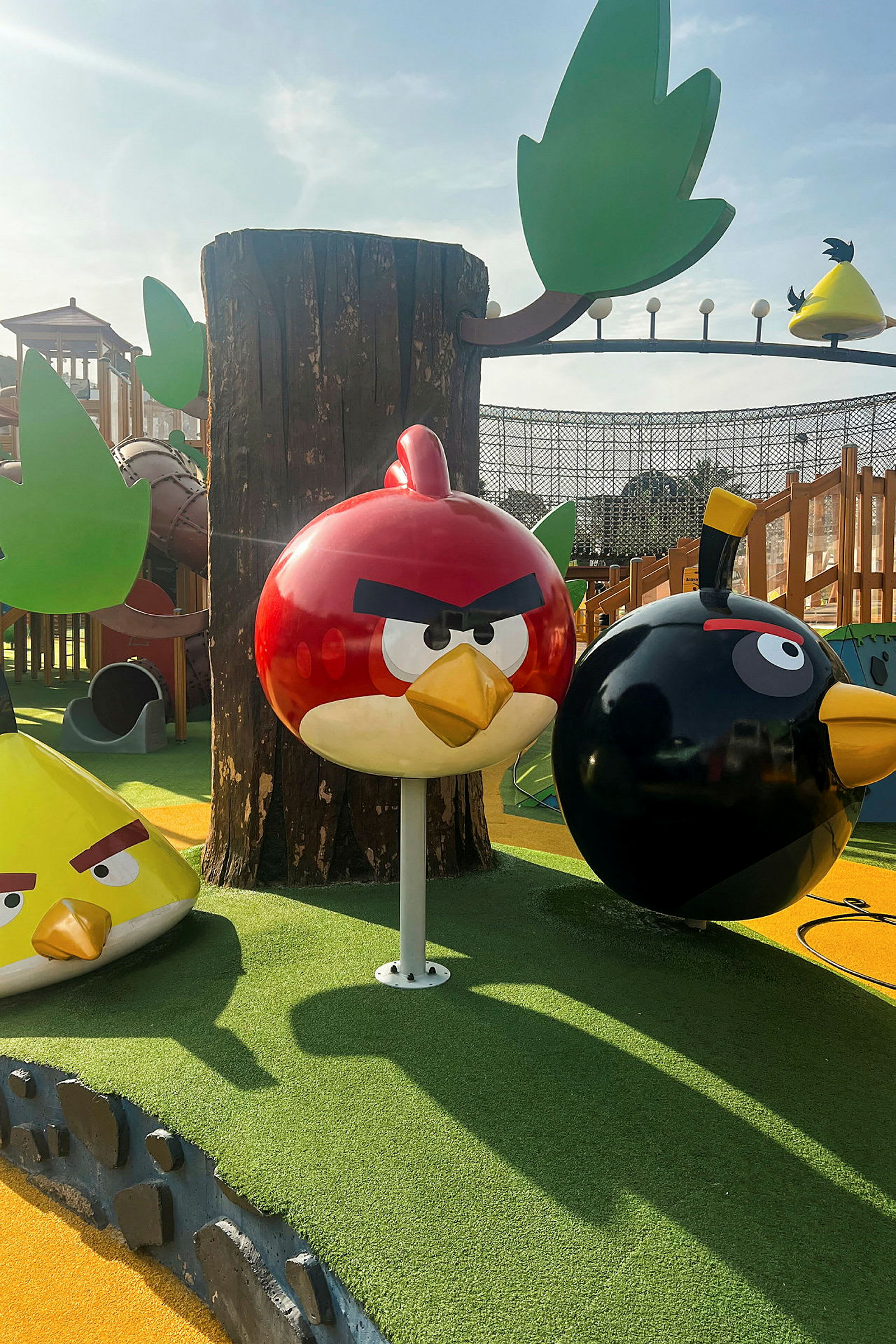 Legeplads med Angry Birds-figurer, inklusive en rød fugl, en sort fugl og en gul trekantet fugl, med træer og andre legestrukturer omkring. (AI-genereret tekst)