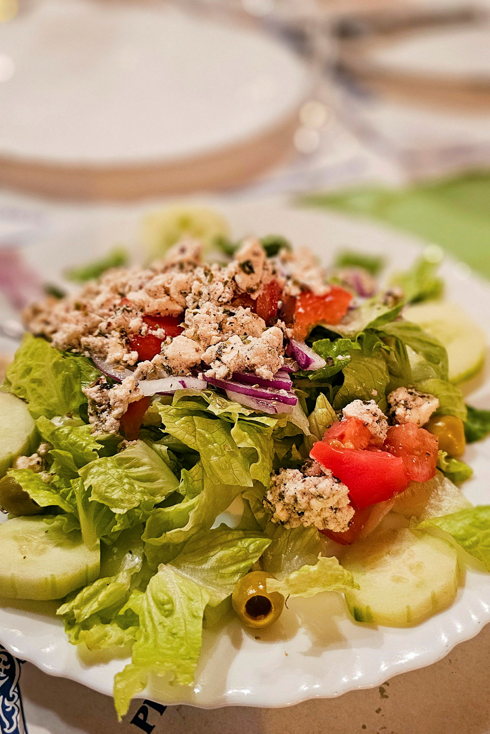 En frisk salat med grønne blade, agurkeskiver, tomatstykker, oliven og smuldret ost på en tallerken. (AI-genereret tekst)