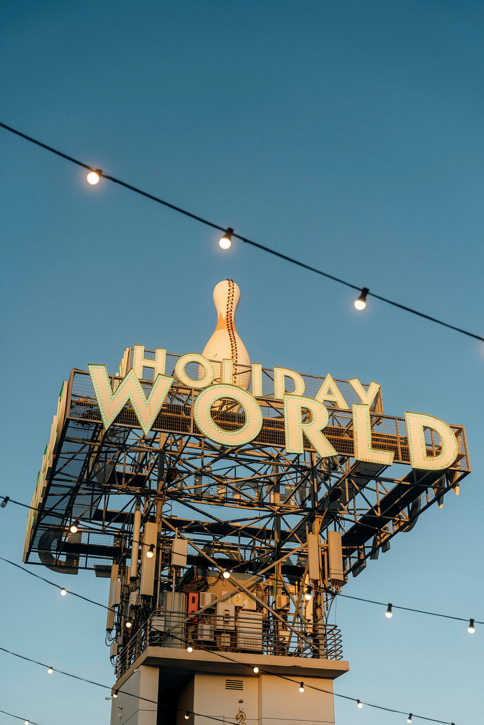 Skiltet med teksten 'Holiday World' og en stor dekoreret bowlingkegle på toppen, med lyskæder mod en klar blå himmel. (AI-genereret tekst)
