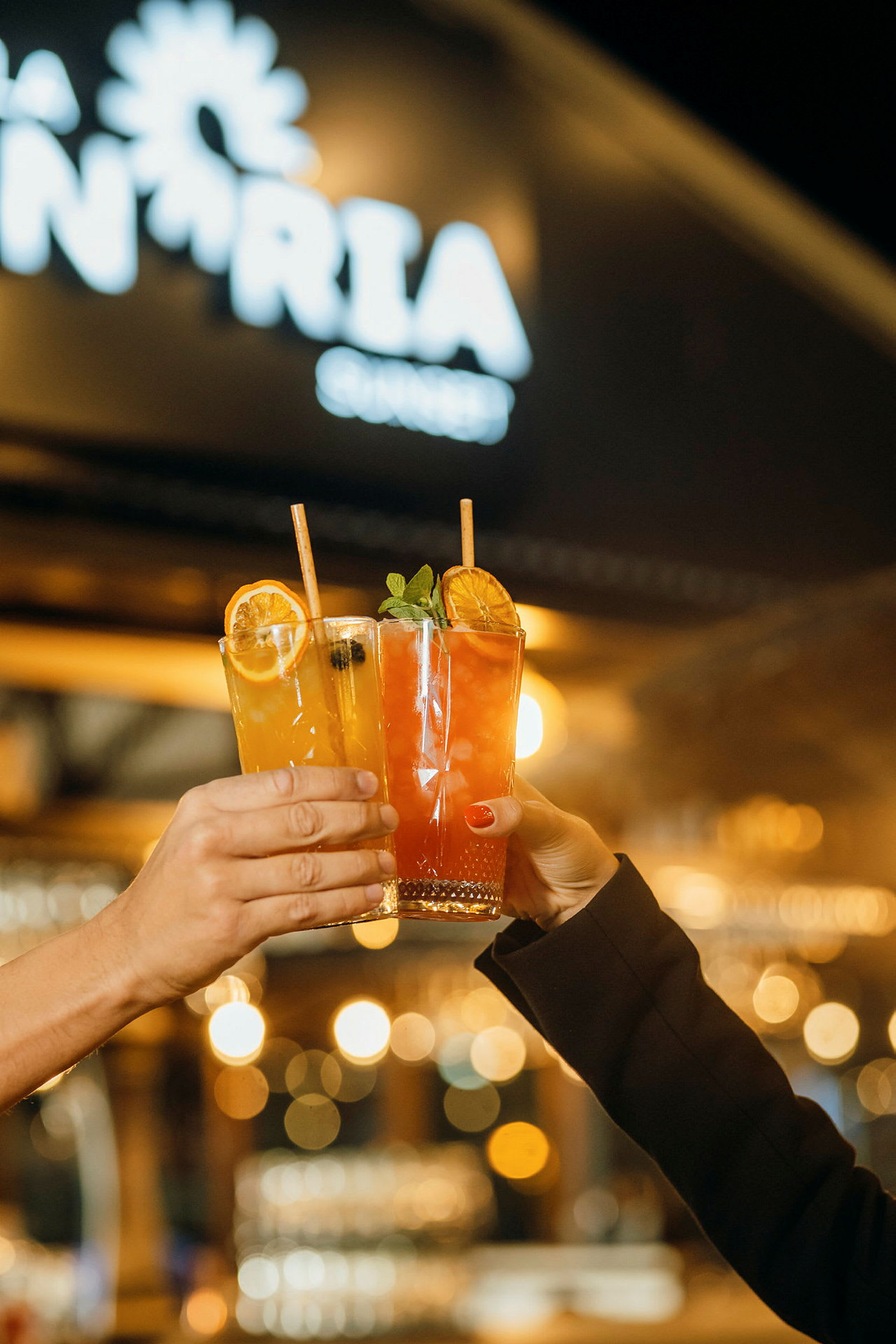 To hænder holder hver deres glas med farverige drinks foran et lysende skilt. (AI-genereret tekst)