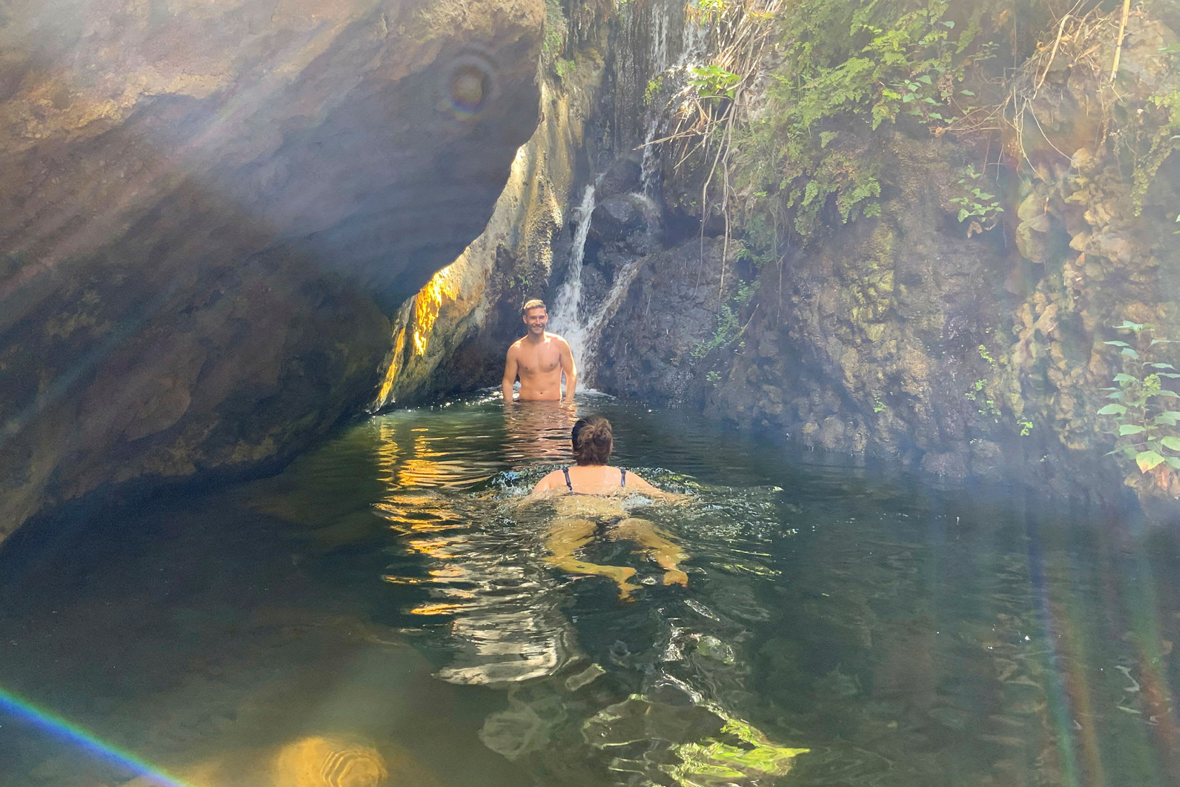 En person svømmer i en naturlig pool nær et lille vandfald omgivet af klipper og vegetation. (AI-genereret tekst)