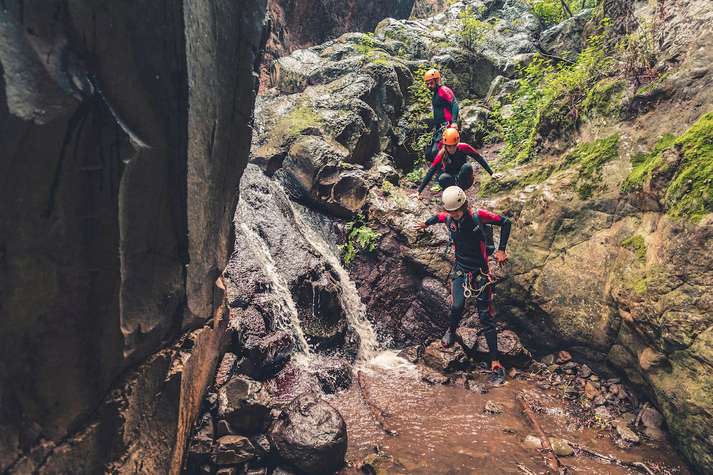Tre personer i klatreudstyr følger en vandstrøm i en klippefyldt ravine. (AI-genereret tekst)
