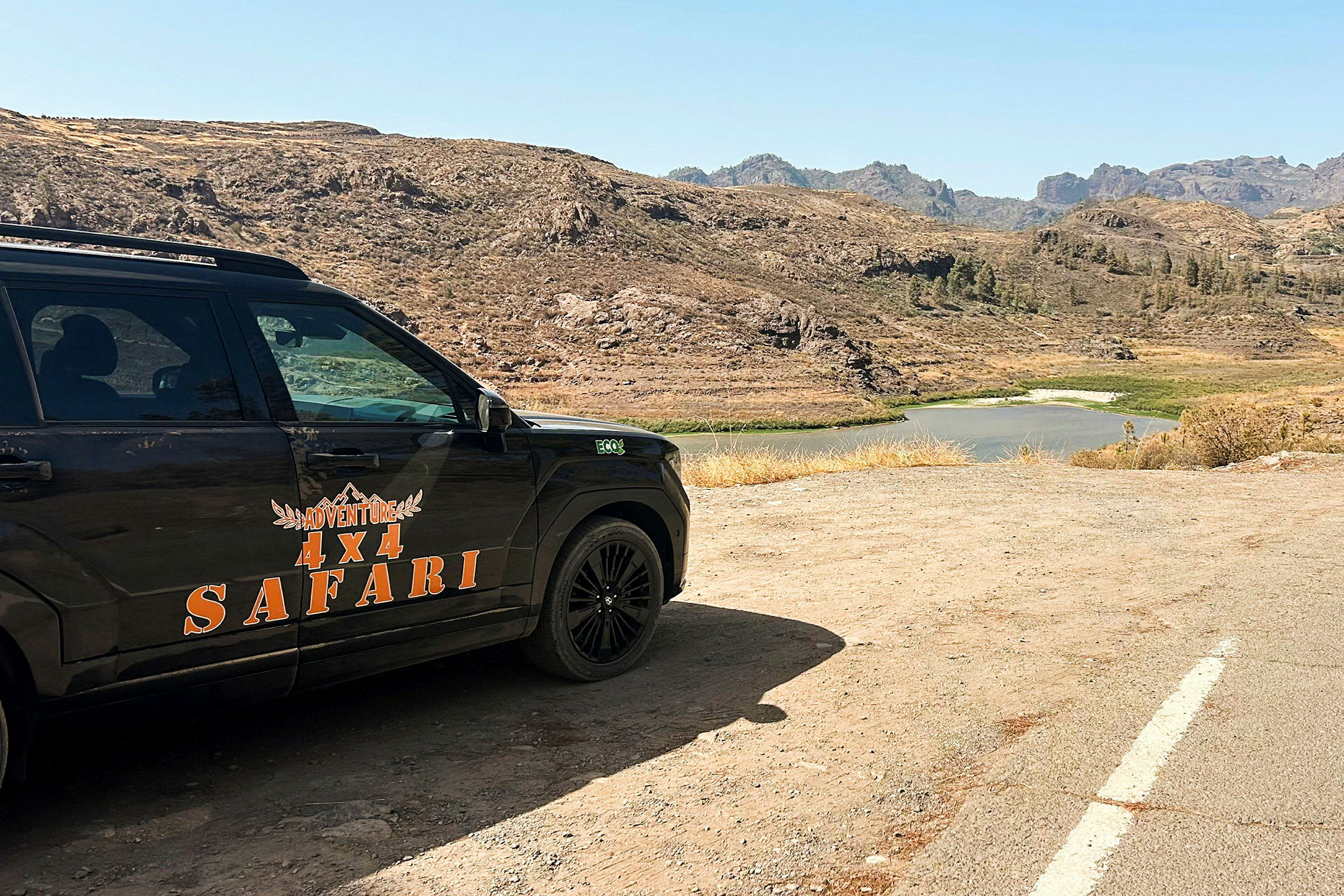 En firehjulstrukket bil med teksten 'Adventure 4x4 Safari' parkeret i et ørkenlignende landskab med en sø i baggrunden. (AI-genereret tekst)