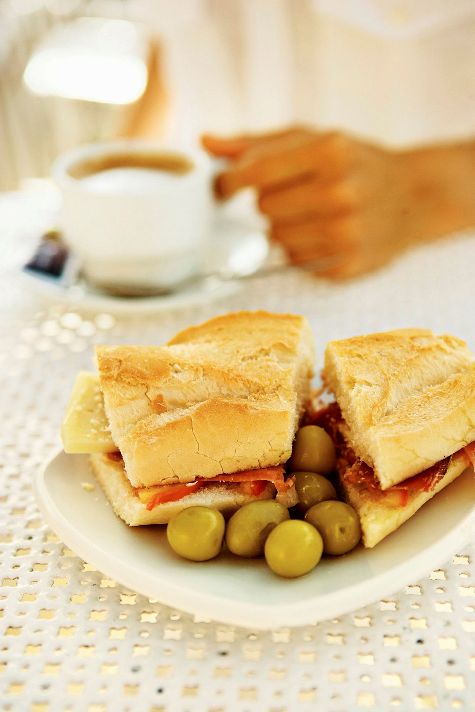 En tallerken med en baguette fyldt med ost og skinke samt nogle oliven. I baggrunden ses en hånd, der holder en kop kaffe. (AI-genereret tekst)