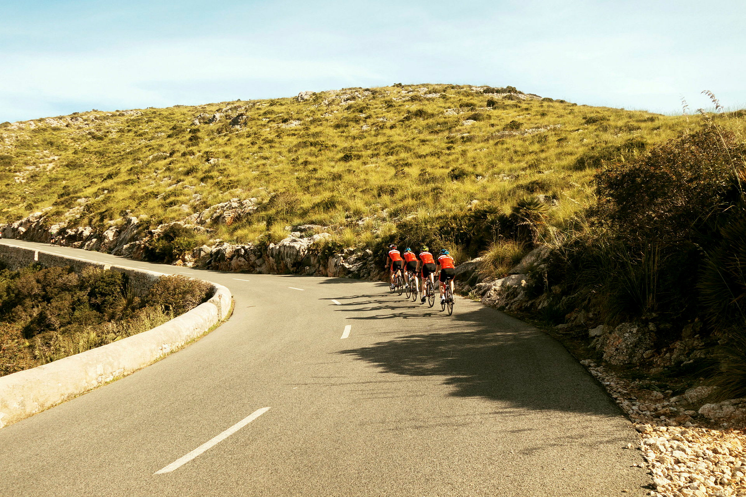 Cykelgruppe på asfaltvej gennem landskab med grøn vegetation og bakker. (AI-genereret tekst)