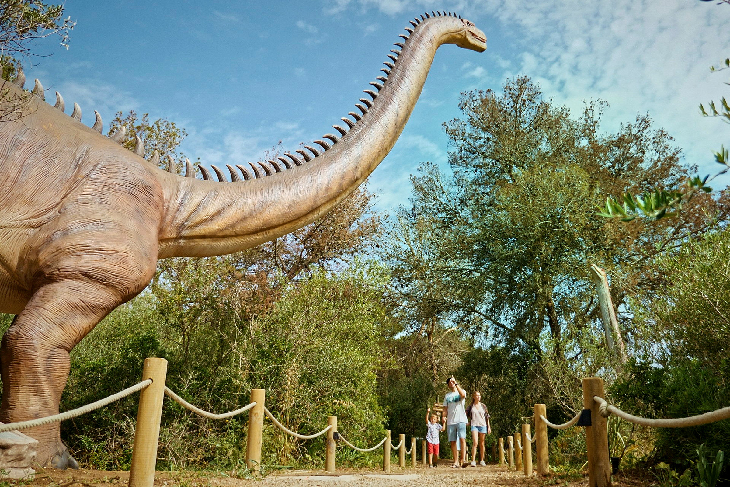 En familie går på en sti ved siden af en stor dinosaurattrap i en udendørs udstilling. (AI-genereret tekst)