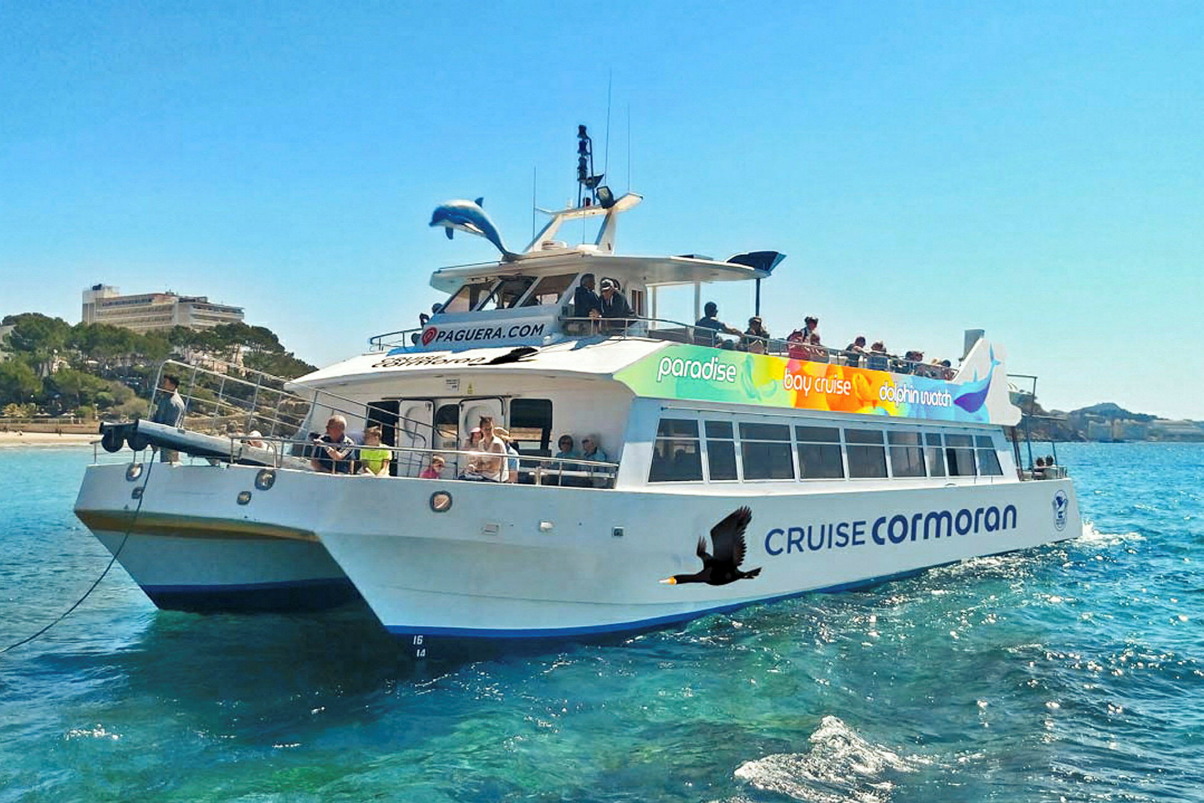 Et skib med navnet 'Cruise Cormoran' på vandet, omgivet af passagerer og med blå himmel og landskab i baggrunden. (AI-genereret tekst)