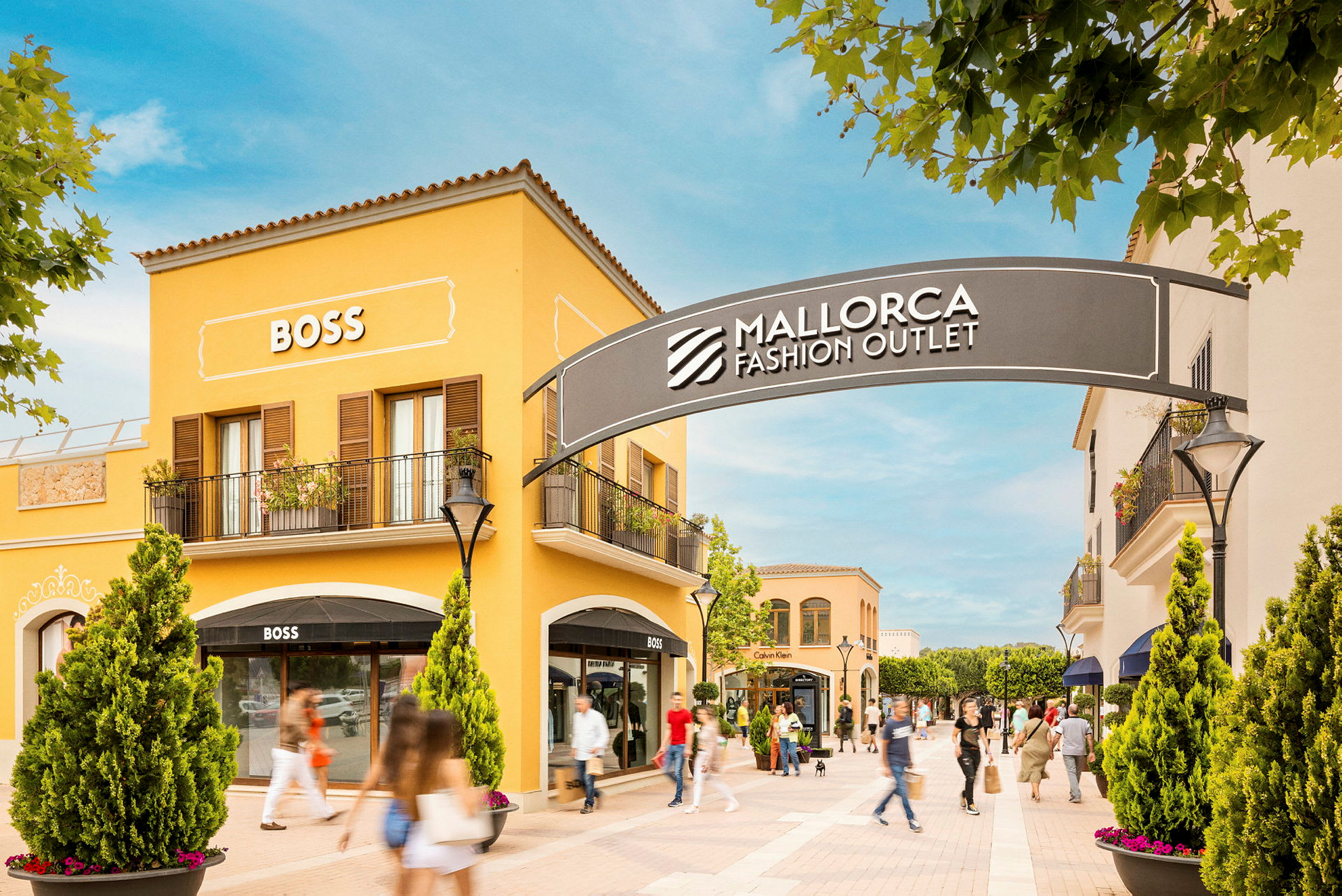 En shoppinggade med gule bygninger, der viser skilte for butikker, herunder BOSS. Et skilt med teksten "Mallorca Fashion Outlet" ses over indgangen til området. (AI-genereret tekst)