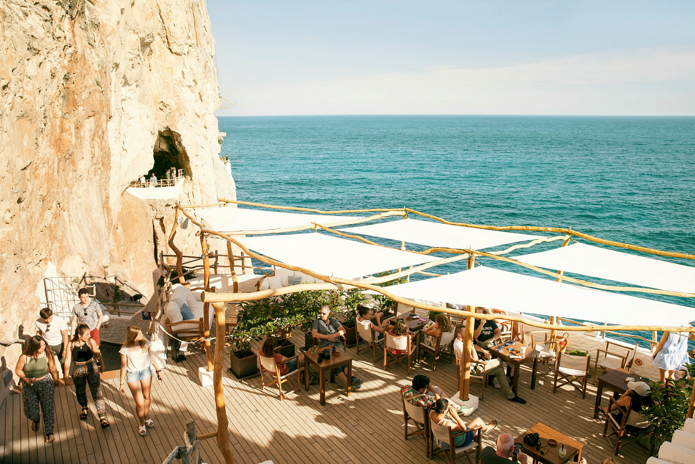En terrasse med borde og stole vendt mod havet, omgivet af klipper. Folk sidder og går på terrassen. (AI-genereret tekst)