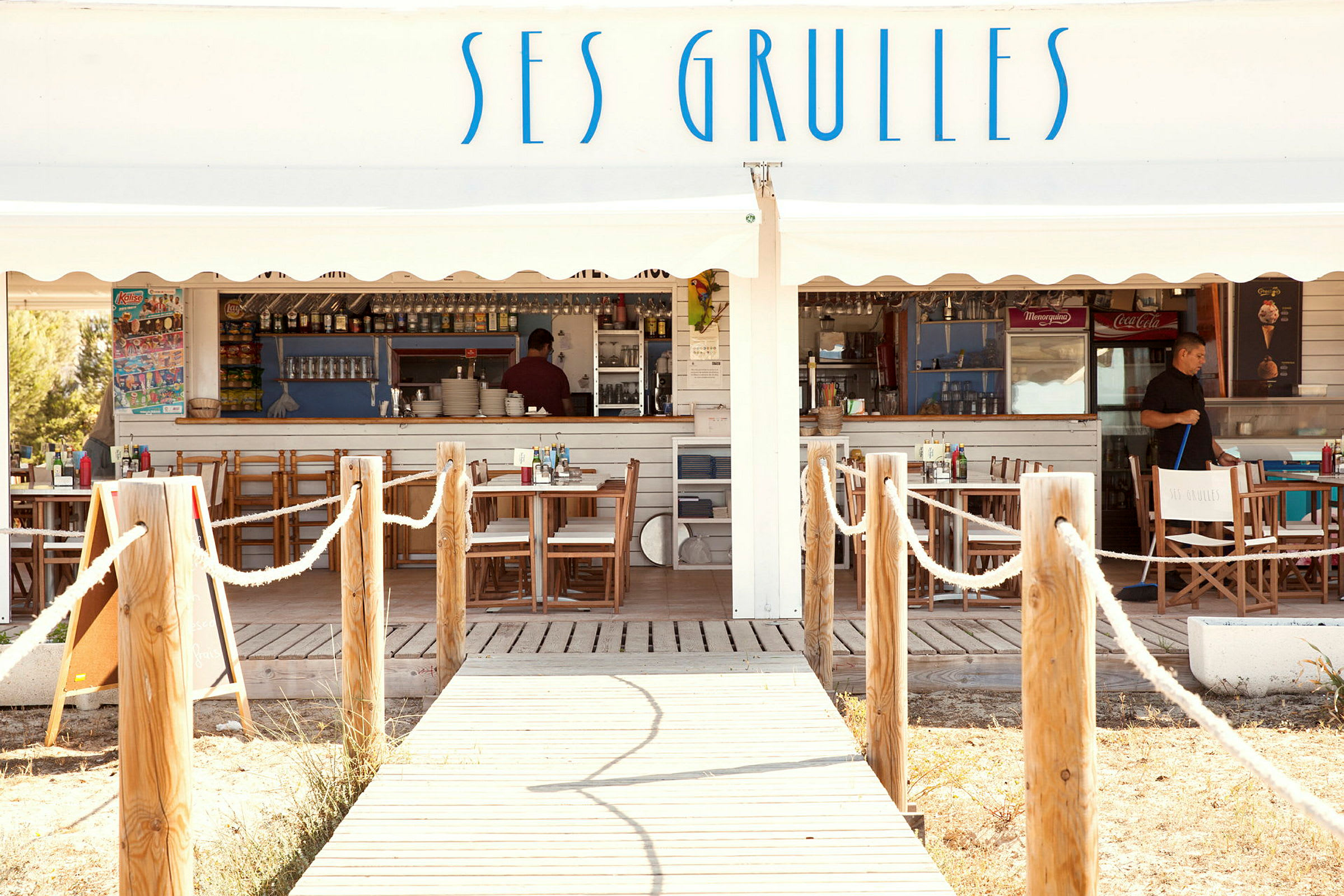 En trægangsti fører til en udendørsrestaurant med hvide trævægge og skiltet "Ses Grulles". (AI-genereret tekst)