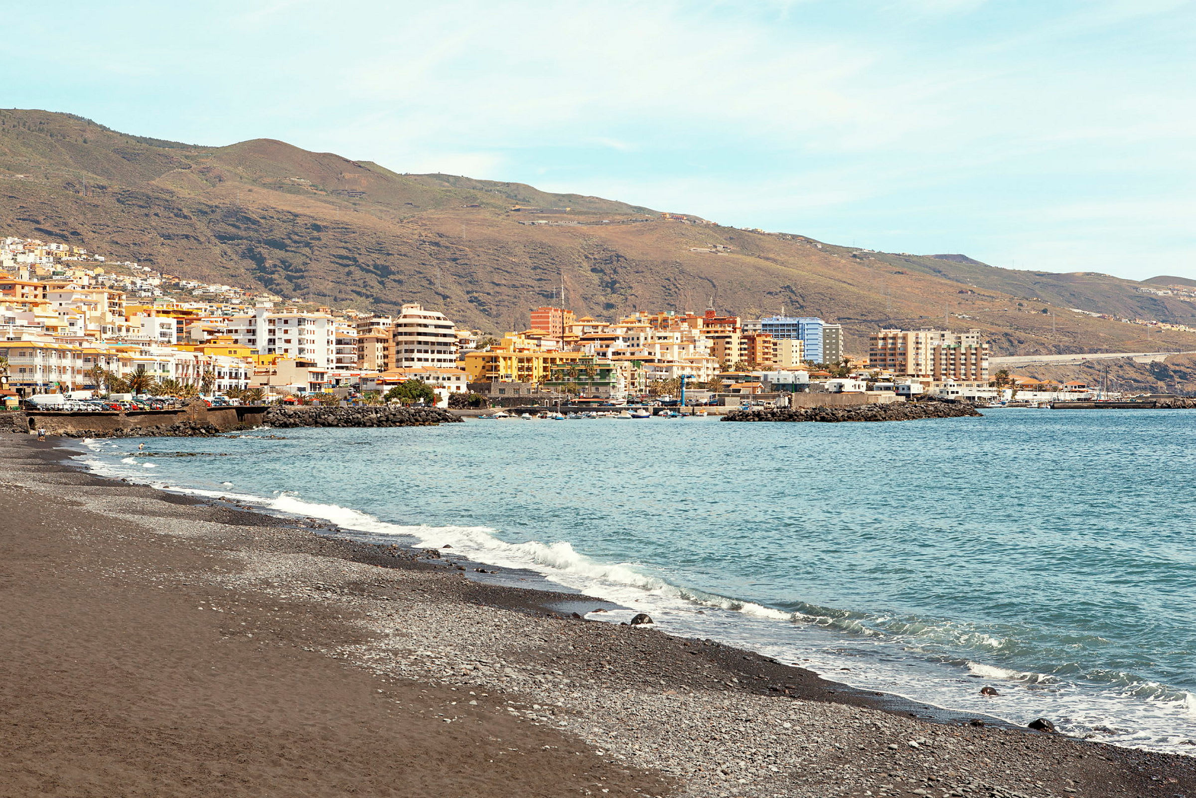 Udsigt over en kyst med sort sandstrand, blåt vand og farverige bygninger ved bjergene i baggrunden. (AI-genereret tekst)