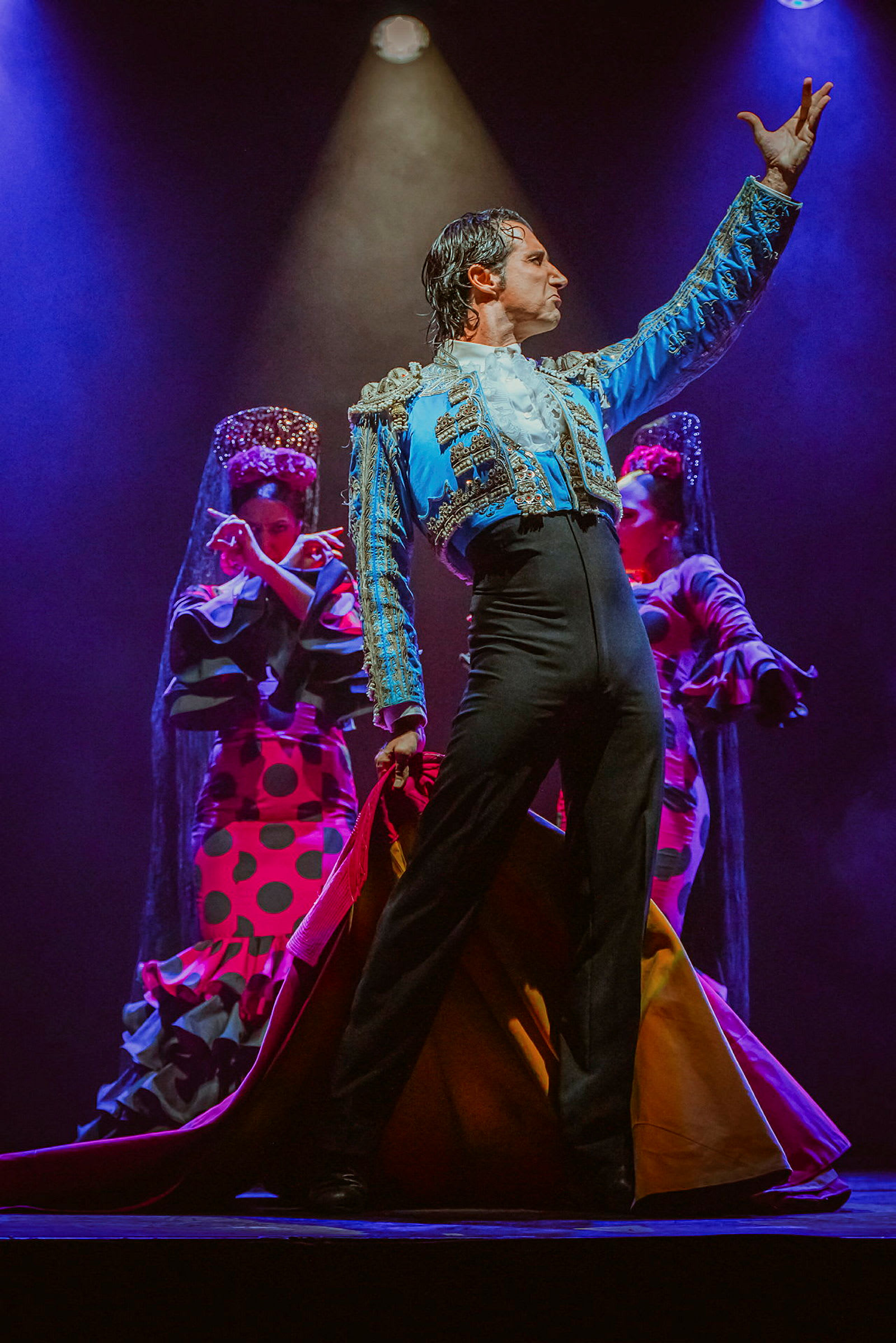 En flamencodanser i farverigt tøj poserer på en scene med to baggrundsdansere i traditionelle dragter. (AI-genereret tekst)