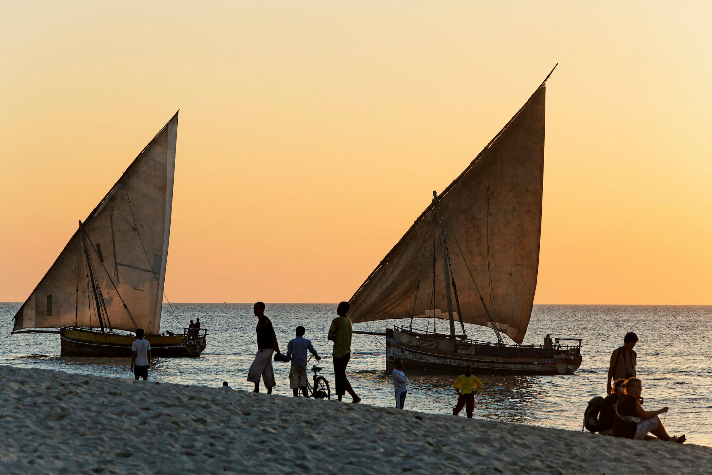 Mennesker går på en strand ved solnedgang. To træbåde med sejl ligger ved vandet. (AI-genereret tekst)