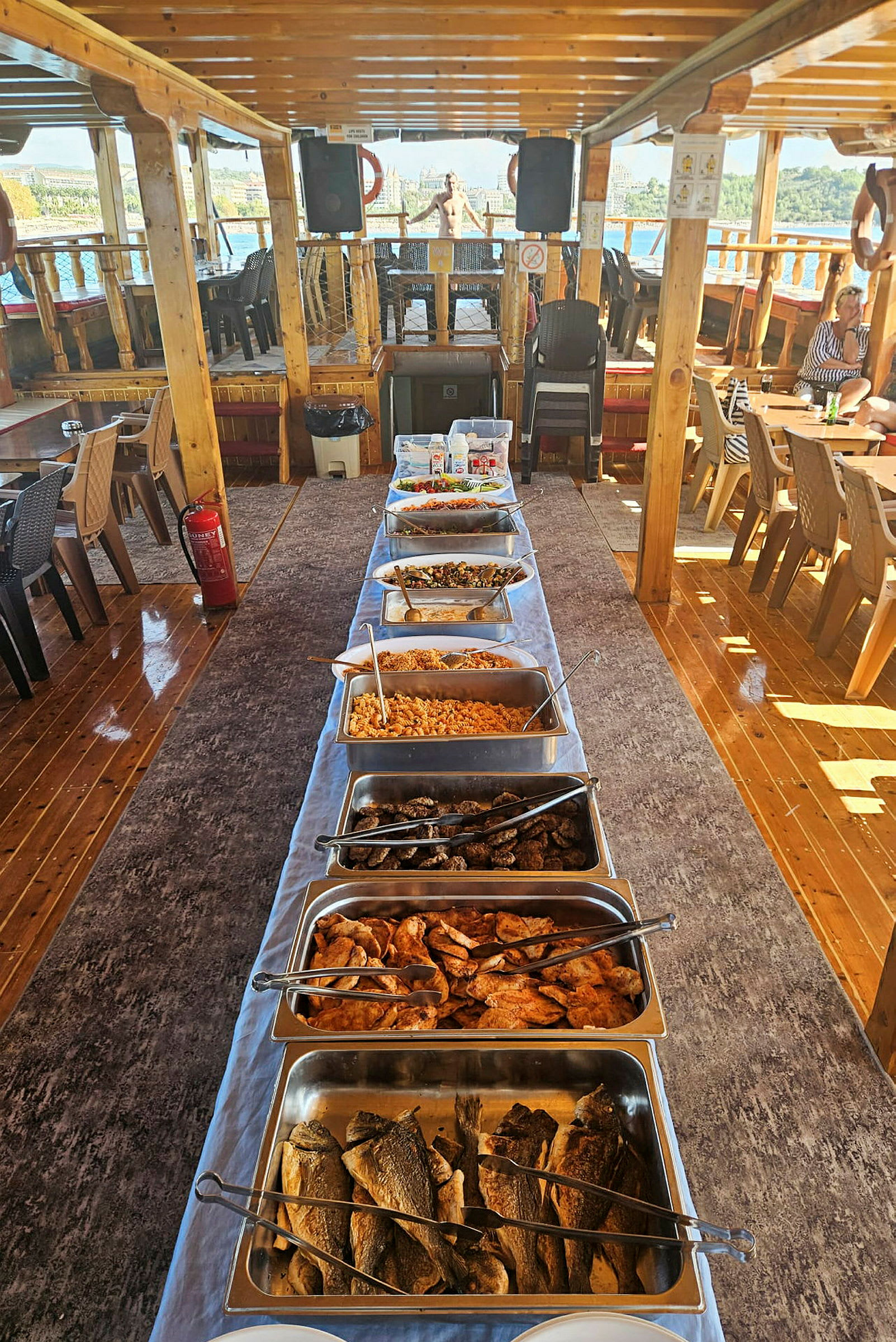 En buffet med forskellige retter er opstillet på en lang række på et overdækket skibsdæk. (AI-genereret tekst)