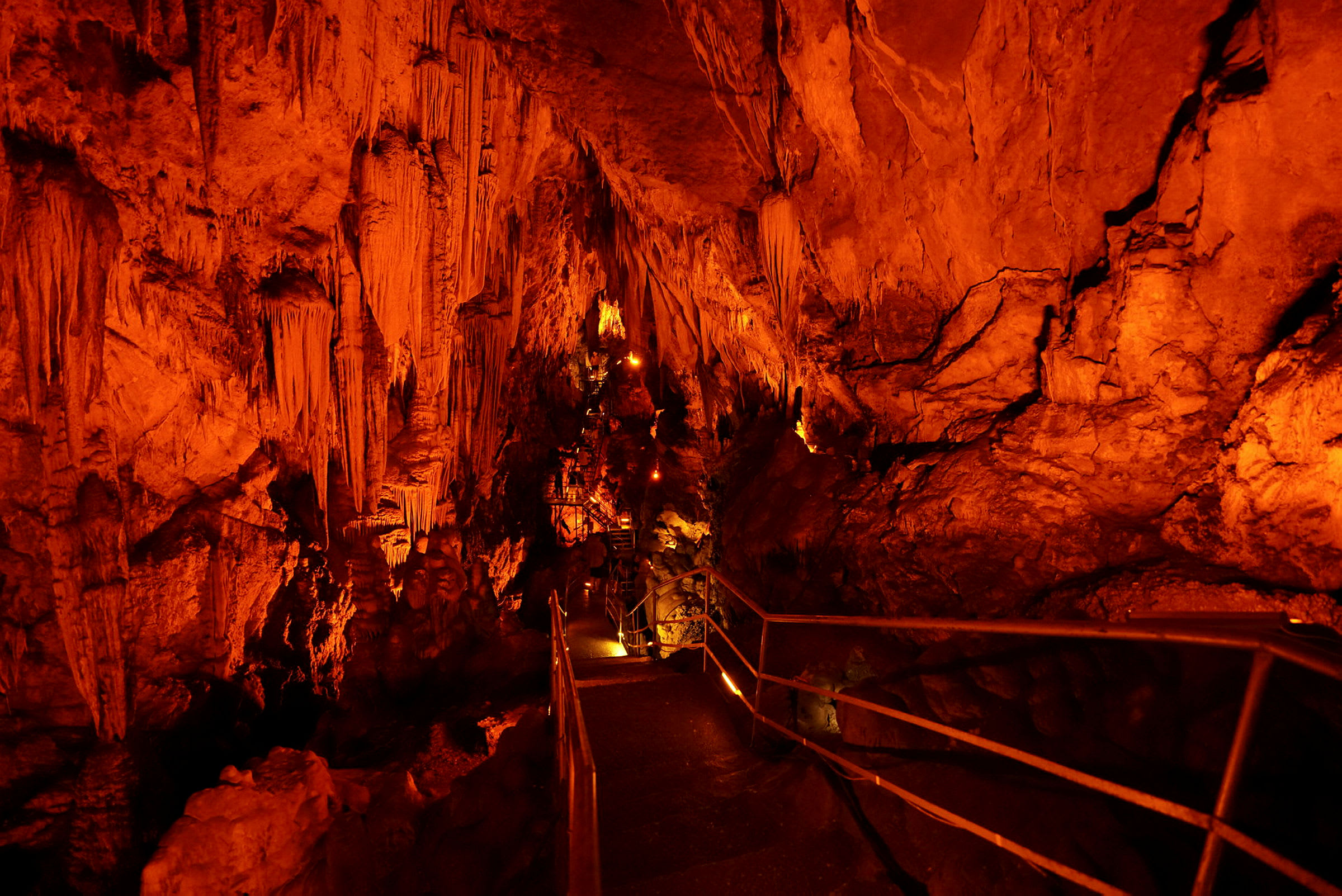 En grotte med stalaktitter og stalagmitter, oplyst med orange belysning. En gangsti med rækværk fører gennem grotten. (AI-genereret tekst)