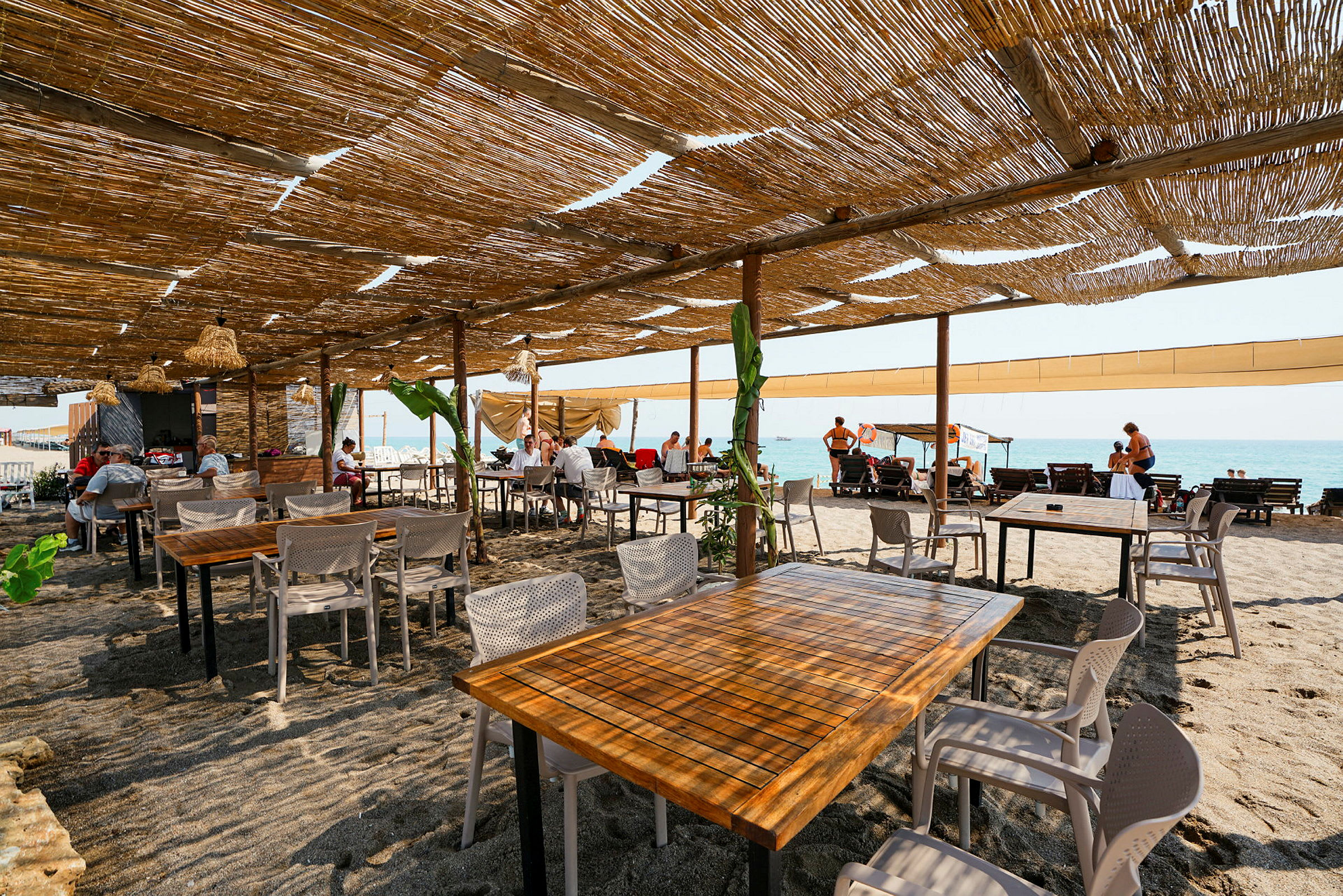 Udendørs restaurant med borde og stole på sandstrand under bambustag. (AI-genereret tekst)