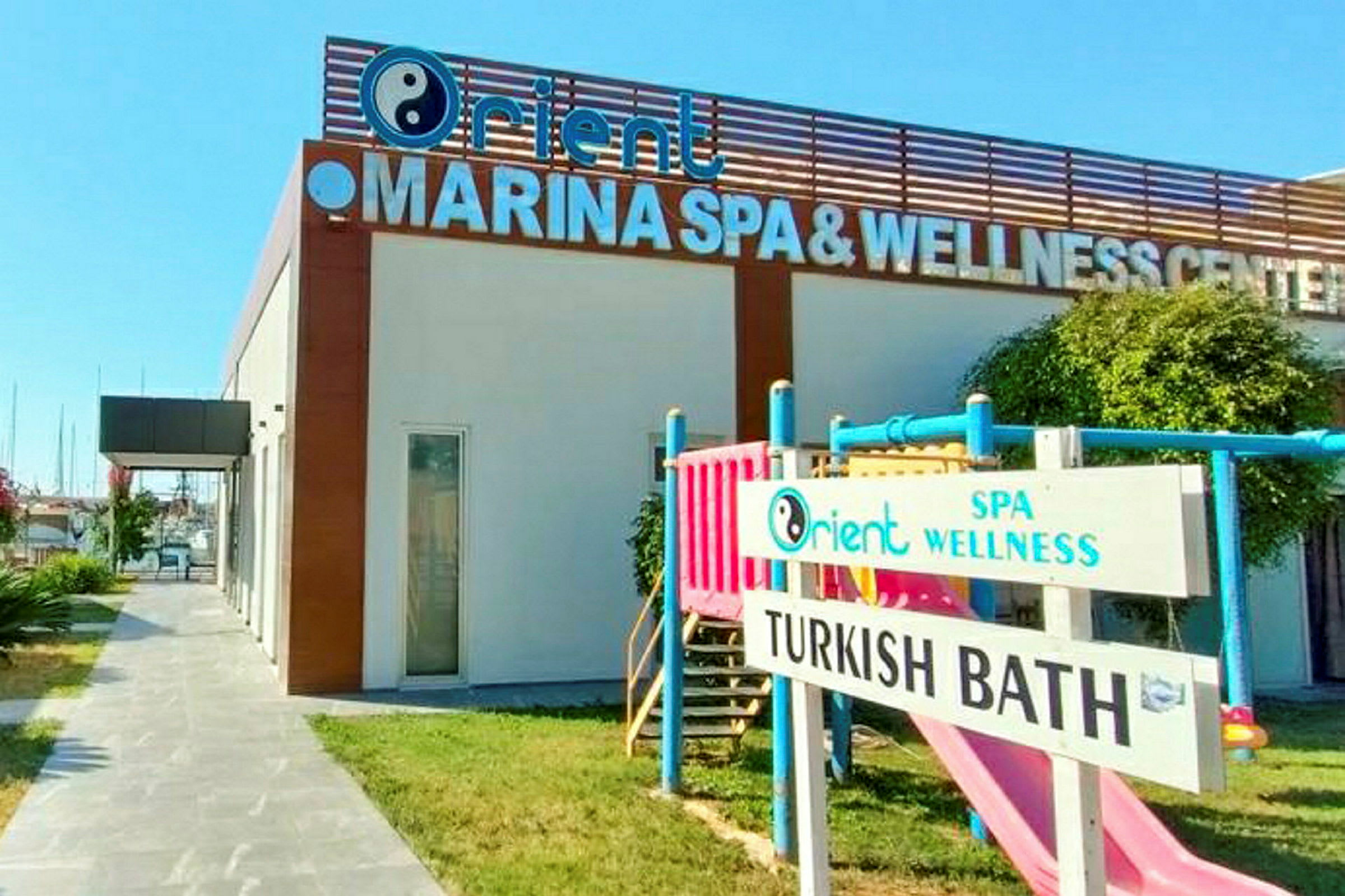 Bygning med et skilt, hvor der står 'MARINA SPA & WELLNESS CENTER'. På græsplænen er der også en legeplads og et skilt med teksten 'TURKISH BATH'. (AI-genereret tekst)