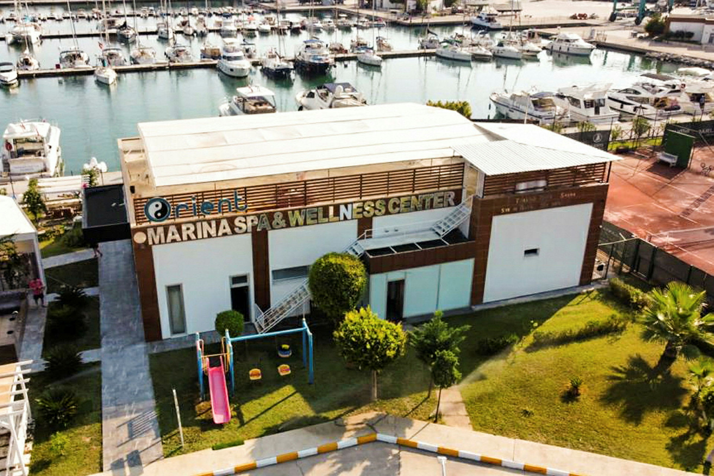 Bygning med skiltet 'Marina Spa & Wellness Center', legeplads med rutsjebane og baggrund med lystbådehavn. (AI-genereret tekst)