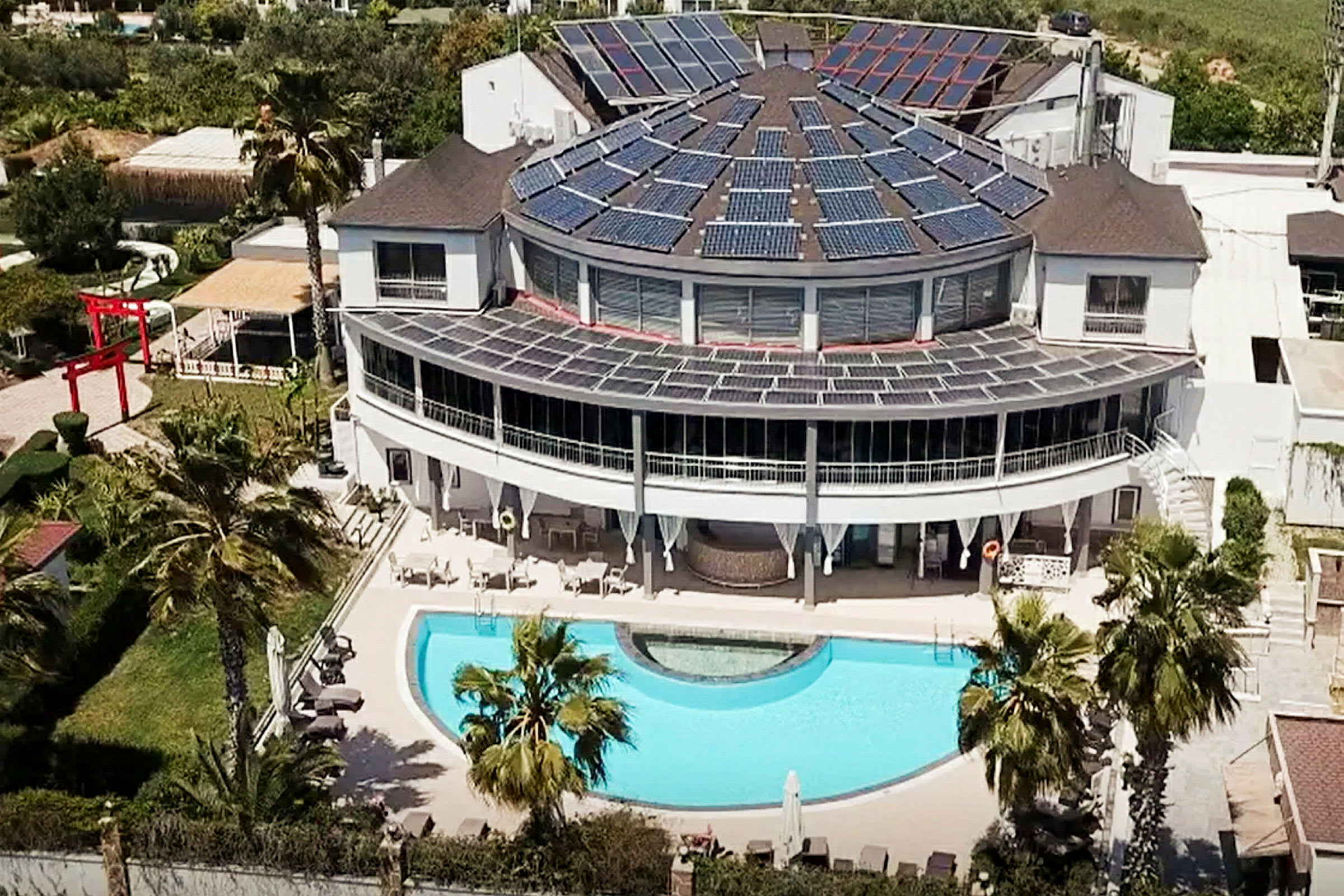 En moderne bygning med solceller på taget og en swimmingpool foran, omgivet af grønne områder. (AI-genereret tekst)