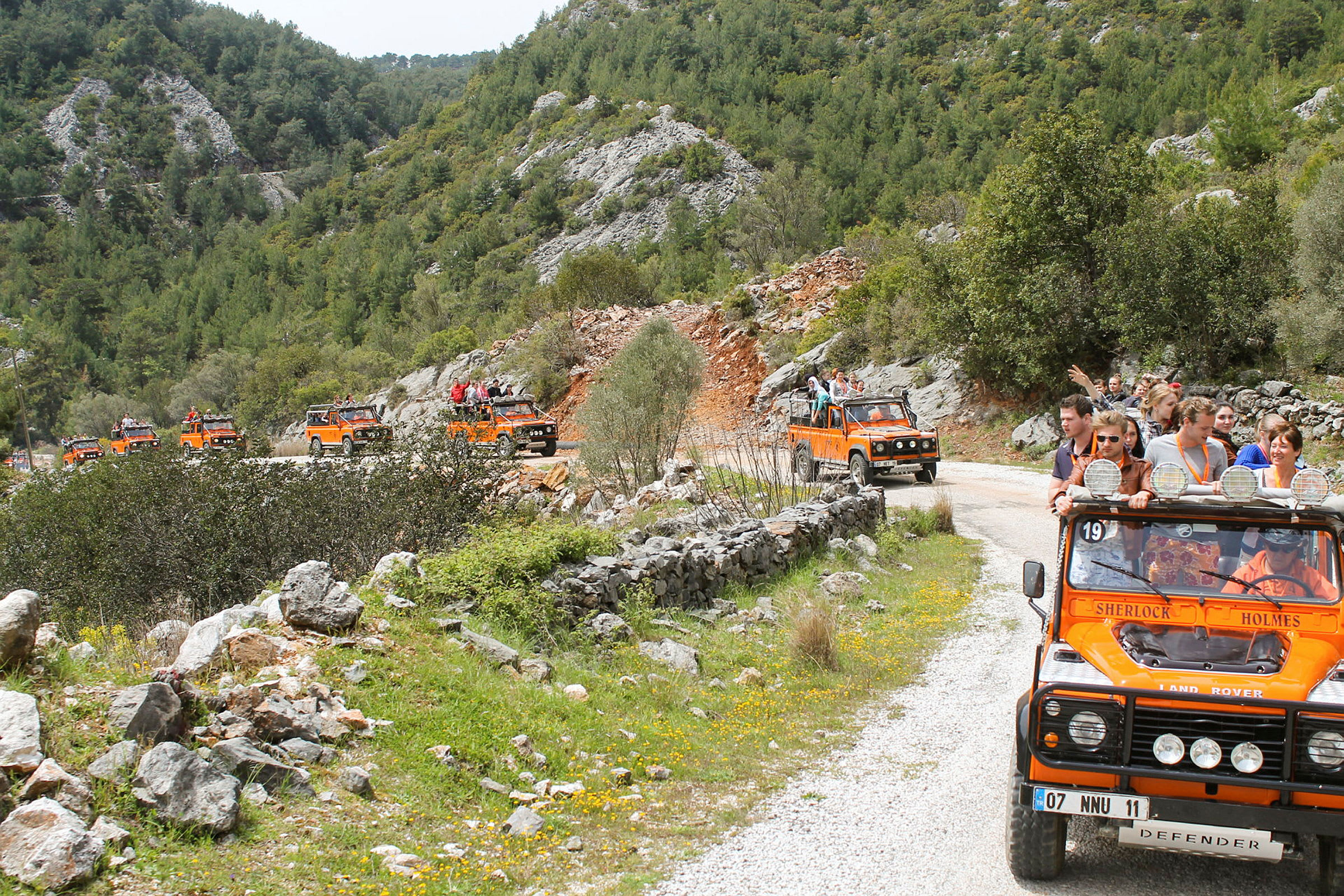 En gruppe orange jeeps kører på en grusvej gennem et bjergrigt og frodigt landskab. (AI-genereret tekst)