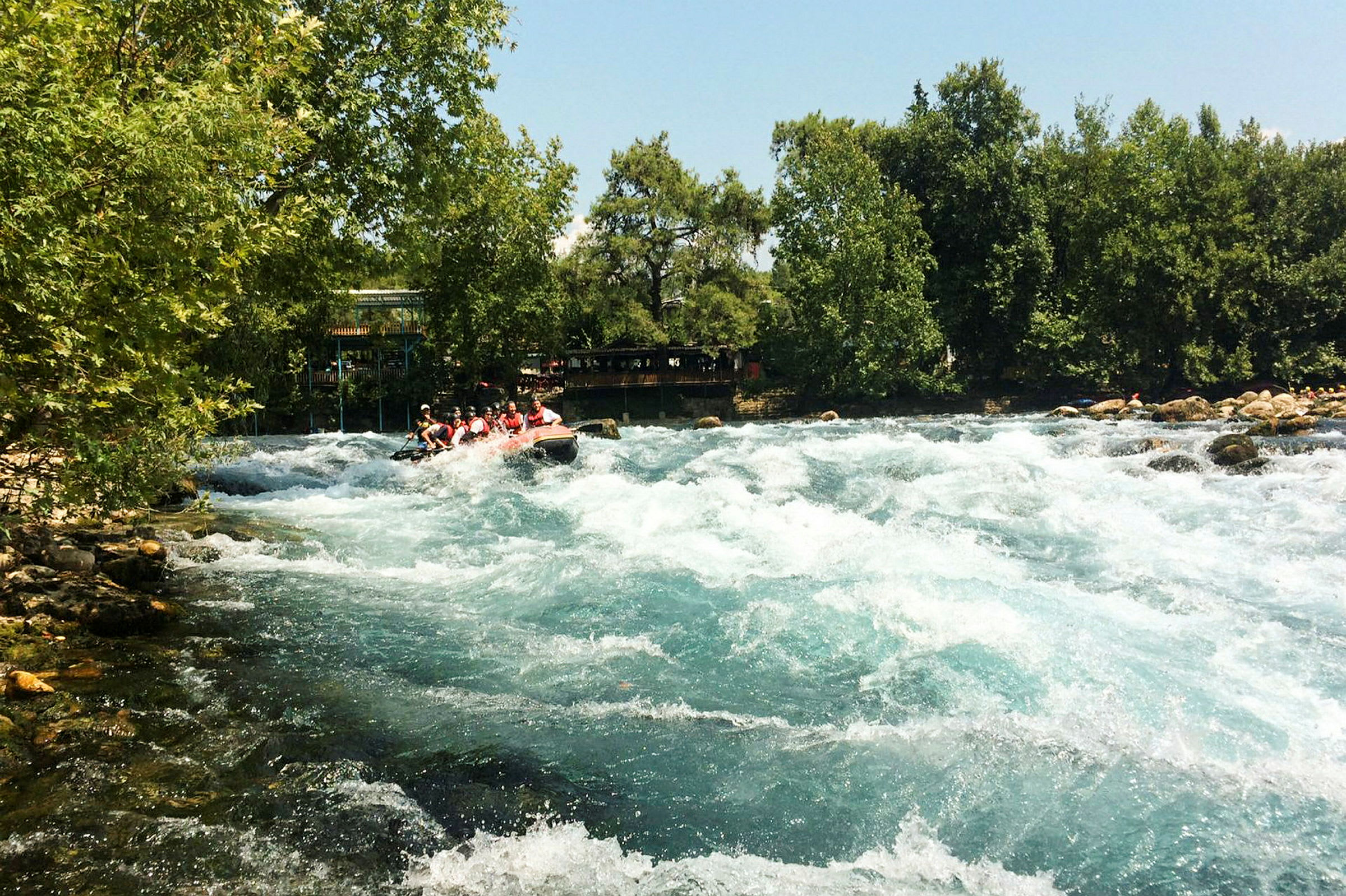 En strømmende flod omgivet af grønt og træer med en oppustelig båd fyldt med personer der driver med riverrafting. (AI-genereret tekst)